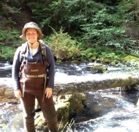 Mindi stands by a river, smiling, wearing a hat and waders